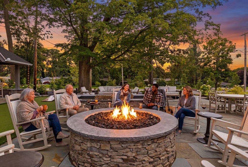 Six adults sit in rocking chairs around a circular stone fire pit on a patio at sunset, surrounded by trees and outdoor furniture at Kilbourn Place on East in Pittsford New York.