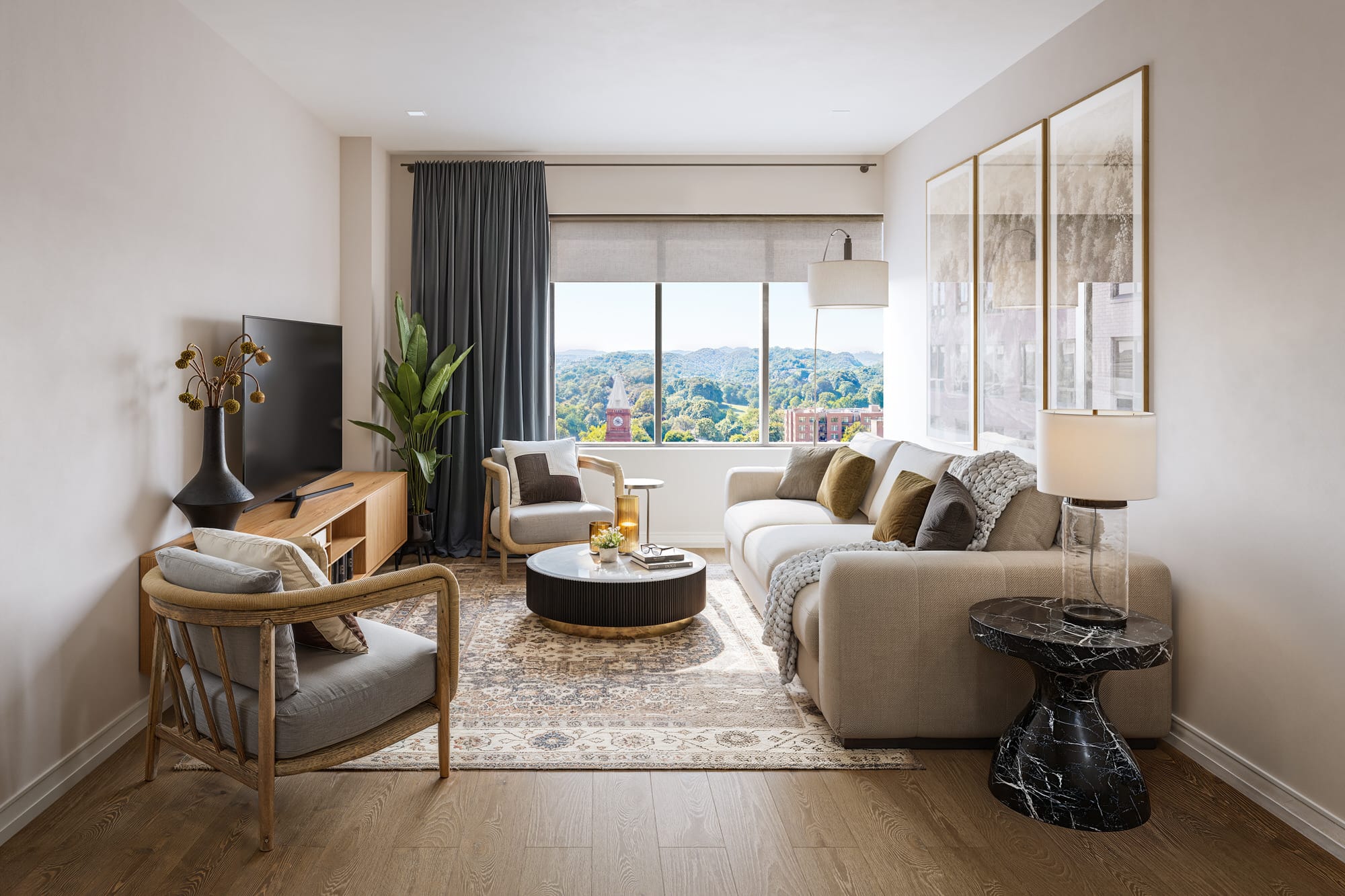 Modern living room with a large window, beige sofa, armchairs, TV on a wooden stand, round coffee table, patterned rug, and framed art on the wall. Cityscape visible through the window.