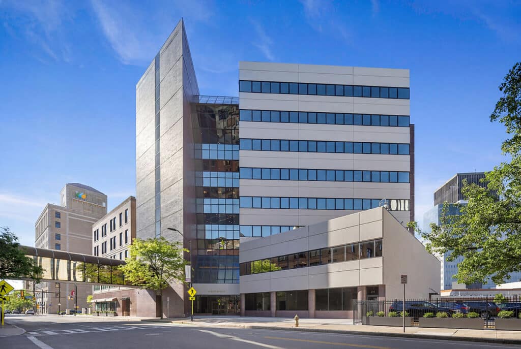 A modern multi-story office building offering premium commercial office space stands on a city street, connected to another building by a skywalk under a clear blue sky.