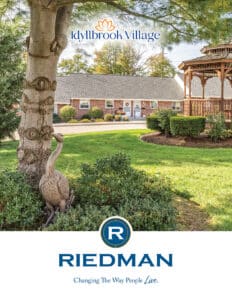 A landscaped yard with a tree, bird statue, and gazebo in front of a brick building. “Idyllbrook Village” and “Riedman” logos appear with the tagline “Changing The Way People Live.”.